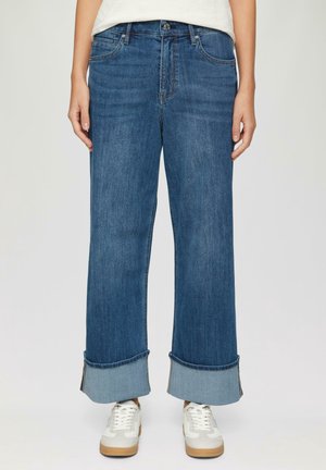 Pohled zepředu na osobu nosící modré džíny s širokými nohavicemi a velkými obrácenými manžetami a bílé tenisky, stojící před jednobarevným pozadím.