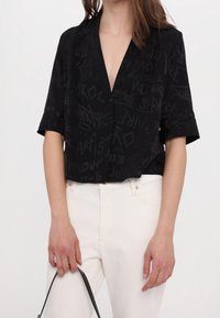 Schwarze Bluse mit kurzen Ärmeln und tiefem V-Ausschnitt, mit einem dezenten Schriftmuster. Kombiniert mit weißen Hosen und einer schwarzen Handtasche.