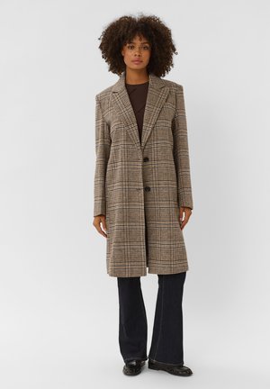 Femme aux cheveux bouclés portant un long manteau marron à carreaux, un haut foncé, un pantalon large noir et des chaussures noires, debout devant un fond uni.