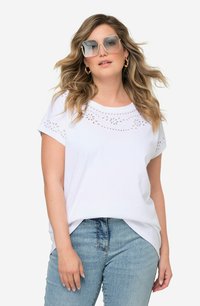 Camiseta blanca de manga corta con detalles de ojal cerca del escote y las mangas, combinada con jeans de mezclilla azules claros. Gafas de sol transparentes y de gran tamaño.