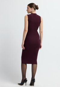 Bordowa, prążkowana sukienka midi bez rękawów z falbaniastym dekoltem. Noszona z przezroczystymi czarnymi rajstopami i czarnymi szpilkami.