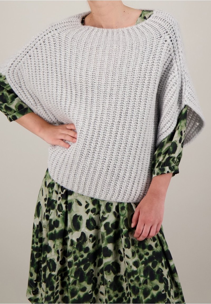 Personne portant un poncho en maille gris clair épais sur une robe à manches longues verte et noire à motifs, une main sur la hanche.
