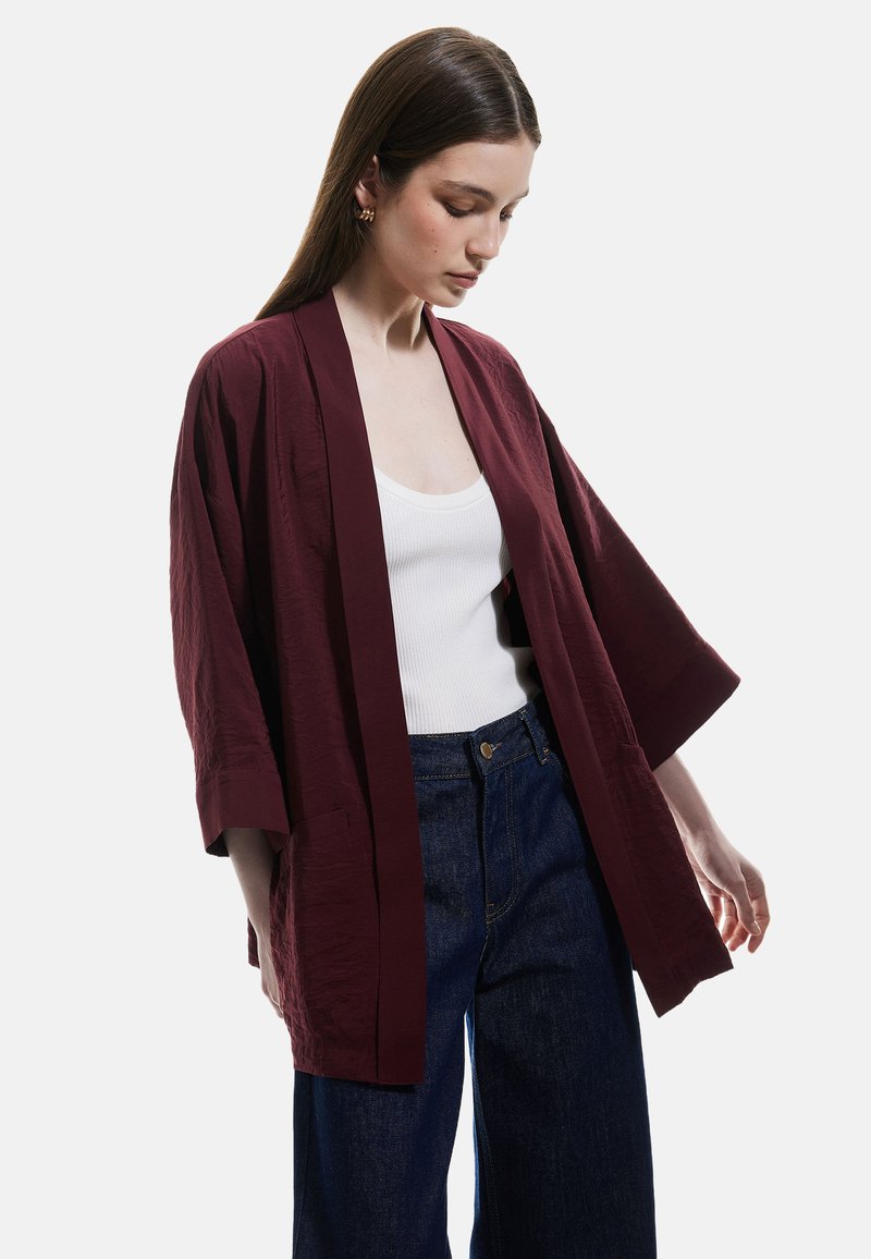 Giacca stile kimono di color bordeaux con maniche larghe, tessuto leggero, apertura frontale e due tasche laterali, indossata sopra una maglietta bianca e jeans scuri.