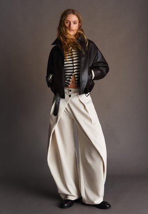 Junge Person trägt eine schwarze Lederjacke, einen gestreiften, bauchfreien Pullover, weite weiße Hosen und schwarze Schuhe, steht vor einem grauen Hintergrund.