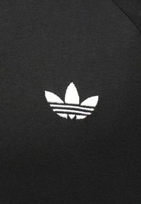 Čierna látka má biele vyšívané logo so tromi listami a tromi horizontálnymi pruhmi na spodnej strane, pričom si zachováva hladkú textúru.