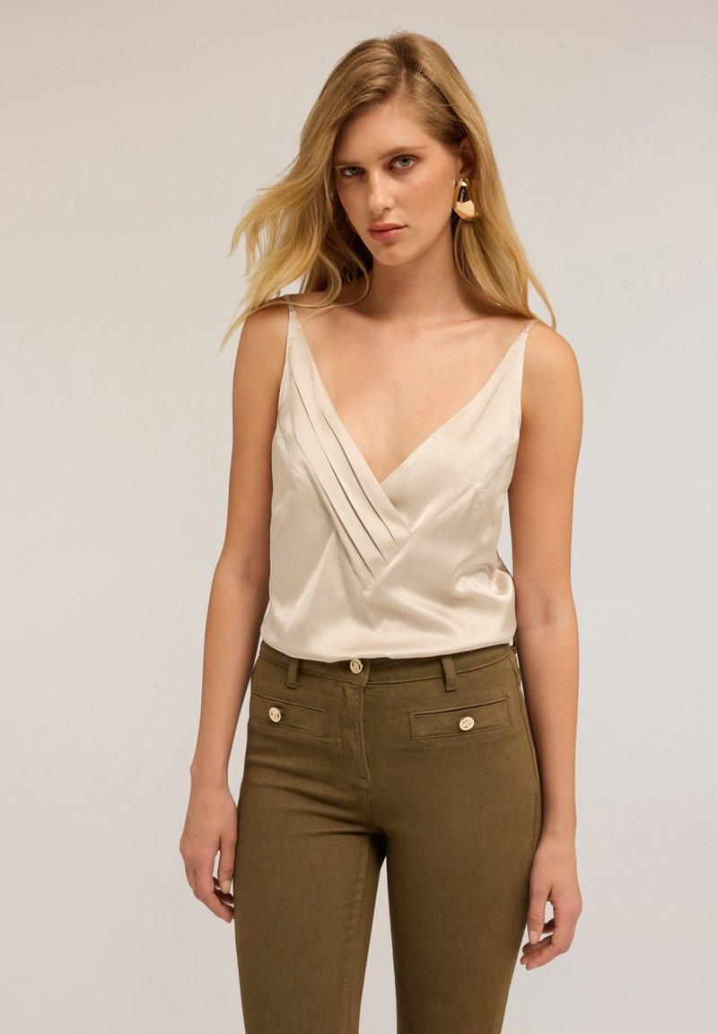Camisole din satin crem, cu decolteu adânc în V și design pliat, asortat cu pantaloni verzi măslinii cu talie înaltă, care prezintă accente din butoane aurii.