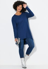 Marineblaue Longsleeve-Tunika mit rundem Halsausschnitt und seitlichen Schlitzen, kombiniert mit dunklen Jeans und metallic-silbernen Ankle Boots. Modell hält eine metallic Clutch.