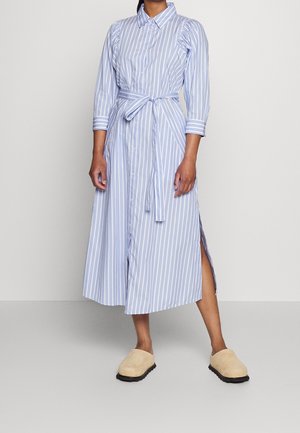 Person iført en midi skjortekjole med blå og hvide lodrette striber, bælte, trekvartlange ærmer, sideslidser og beige slip-on sko.