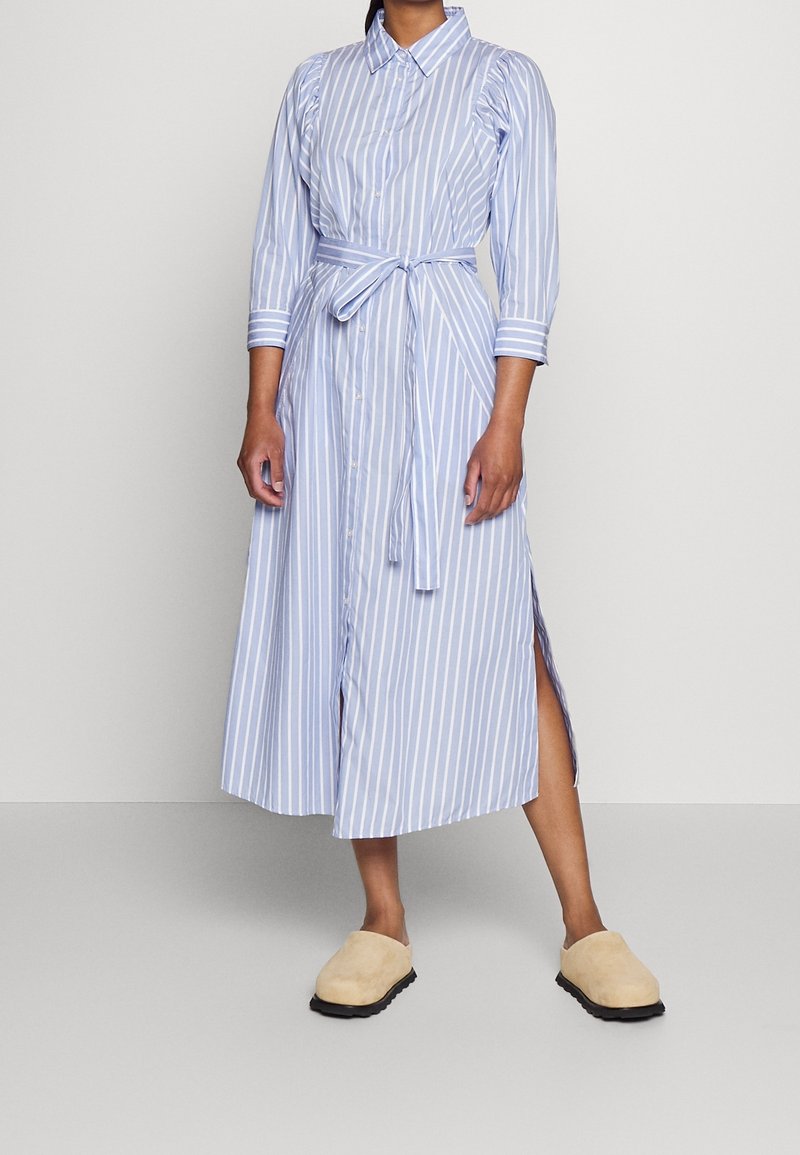 Personne portant une robe chemise midi à rayures verticales bleues et blanches avec ceinture, manches trois-quarts, fentes latérales et chaussures beiges à enfiler.
