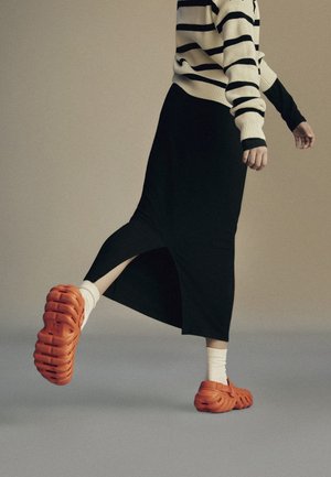 Person in schwarzem Rock mit Seitenschlitz, gestreiftem Pullover, weißen Socken und orangefarbenen Slip-On-Schuhen, die auf neutralem Hintergrund nach vorne tritt.