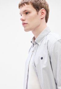 Πουκάμισο με κουμπιά σε ανοιχτό γκρι χρώμα με λεπτές κάθετες ρίγες. Διαθέτει γιακά και κεντημένο λογότυπο στην τσέπη στο στήθος.