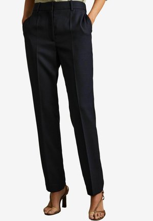 Pantalones oscuros de sarga en navy con piernas rectas, que cuentan con bolsillos laterales y un sutil patrón texturizado, combinados con tacones marrones de tiras.