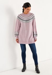 Hellrosa gestrickter Pullover mit Rundhalsausschnitt, gemustertem Einsatz in Grau, Schwarz und Weiß. Kombiniert mit dunkelblauen Jeans und schwarzen Ankle Boots.