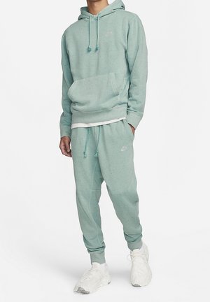 Mężczyzna ubrany w miętowo-zieloną bluzę z kapturem w plamki i dopasowane dresy, w białych sportowych butach, stojący na białym tle.