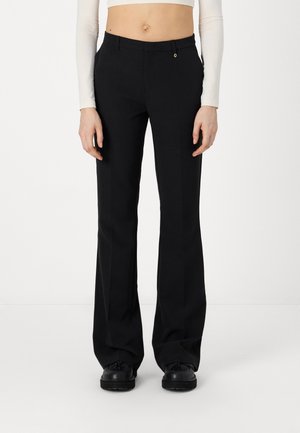 Personne portant un pantalon évasé taille haute noir, des chaussures noires et un top court blanc à manches longues, debout devant un fond uni.