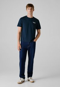 Navyblauwe katoenen T-shirt met korte mouwen en een ronde hals, met een klein wit logo op de linkerkant van de borst. Gdragen met donkere spijkerbroek.