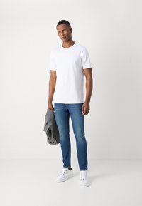 White short-sleeve t-shirt, blue slim-fit jeans, holding a gray jacket. White sneakers with laces, plain design, minimal details.