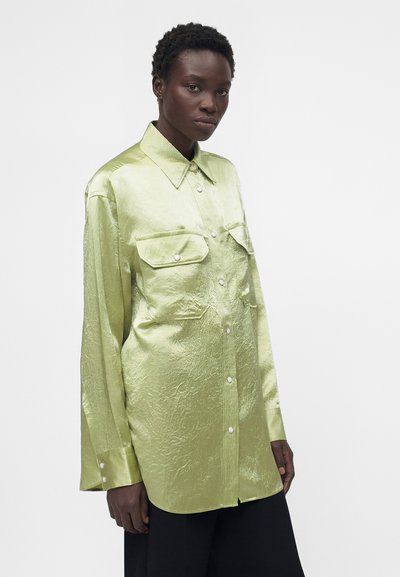 Γυναίκα που φοράει ένα μακρυμάνικο κουμπωτό πουκάμισο σε γυαλιστερό ανοιχτό πράσινο χρώμα με δύο τσέπες στο στήθος, στέκεται μπροστά σε λευκό φόντο.