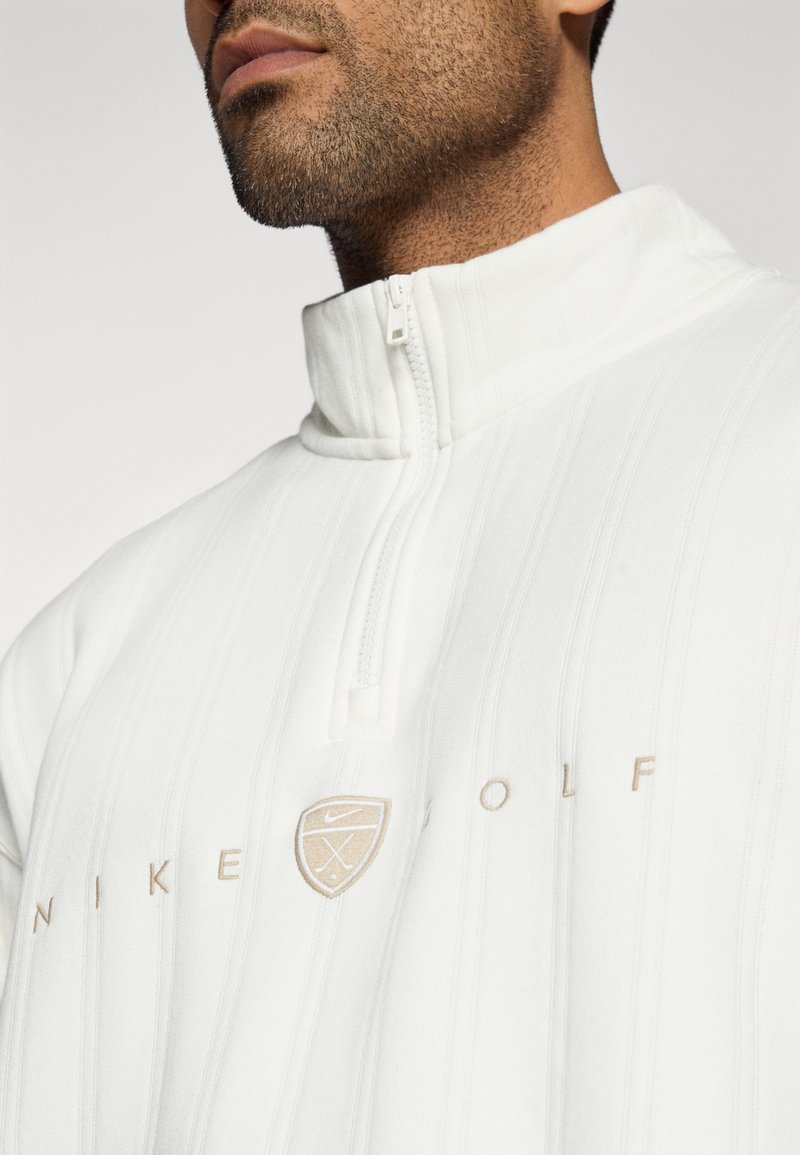 Weißer Nike Golf-Pullover mit hohem Kragen, halber Reißverschluss, gestreiftem Muster und dezentem beigen Branding auf der Brust.