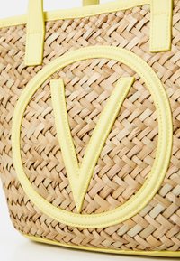 Vävd halm tote-väska med gula läderdetaljer. Har en stor gul "V"-logga och hållbara handtag. Texturen är intrikat och naturlig.