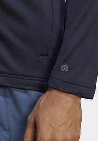 Granatowa koszula z długim rękawem z teksturowanej tkaniny, z małym logo globusa na mankiecie. Czyste liniowe szwy i prosta dzianina.