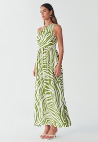 Maxikjole i grøn og hvid zebramønster med ene skulder, markeret talje og flydende stof. Båret med hvide sko med hæl.