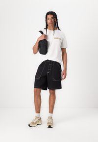 Hvit t-skjorte med gul logo, svarte shorts med hvit søm, og beige sneakers. Modellen bærer en svart crossbody-veske.