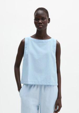 Vrouw met kort haar die een mouwloos lichtblauw topje en bijpassende broek draagt, staand met één hand in de zak tegen een effen witte achtergrond.