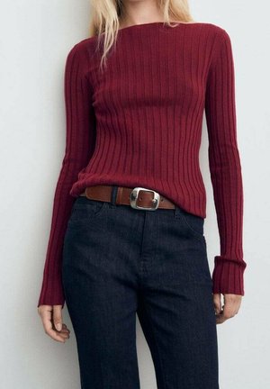 Vrouw draagt een aansluitende bordeauxrode geribde trui met lange mouwen, ingestopt in donkere hoge taille broek met een bruine riem.