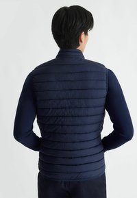 Gilet imbottito blu navy con design a trapunta, chiusura con zip frontale, colletto alto e materiale leggero, indossato sopra una maglietta a maniche lunghe.