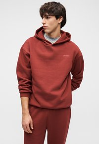 Sweat à capuche bordeaux en tissu doux, avec capuche à cordon et poche kangourou. Coupe décontractée avec poignets côtelés, assorti à un pantalon de survêtement.