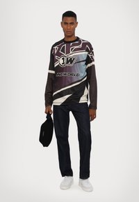 Langärmliges Grafikshirt mit einem Design in Schwarz, Blau und Weiß sowie Text; kombiniert mit dunklen Jeans und weißen Sneakers, hält eine schwarze Tasche.