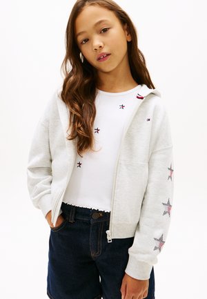 Pige med langt brunt hår iført en hvid lynlåshættejakke med stjernedesign og mørke denimshorts mod en ensartet lys baggrund.