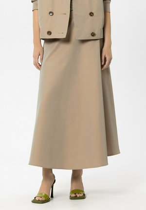 Frau trägt einen beigen, knöchellangen Rock, eine beige Jacke mit großen Knöpfen und olivgrüne, offene High-Heel-Sandalen.