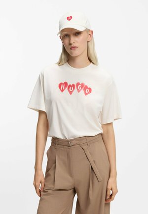 Cream short-sleeve t-shirt with a red heart design and "HUGO" text. Beige tailored pants with a belted waist. White cap with a heart logo.