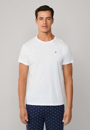 Weißes Baumwoll-T-Shirt mit rundem Halsausschnitt und kurzen Ärmeln, auf der Brust mit einem kleinen dunkelblauen Logo versehen. Kombiniert mit dunkelblauen gemusterten Hosen.