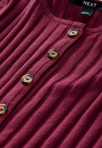 Burgundy ribbad stickad topp med rundad halsringning och knappstängning, prydd med stora kontrasterande knappar och en texturerad tyg.