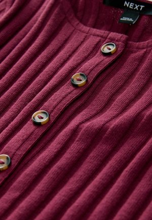 Burgundy ribbad stickad topp med rundad halsringning och knappstängning, prydd med stora kontrasterande knappar och en texturerad tyg.