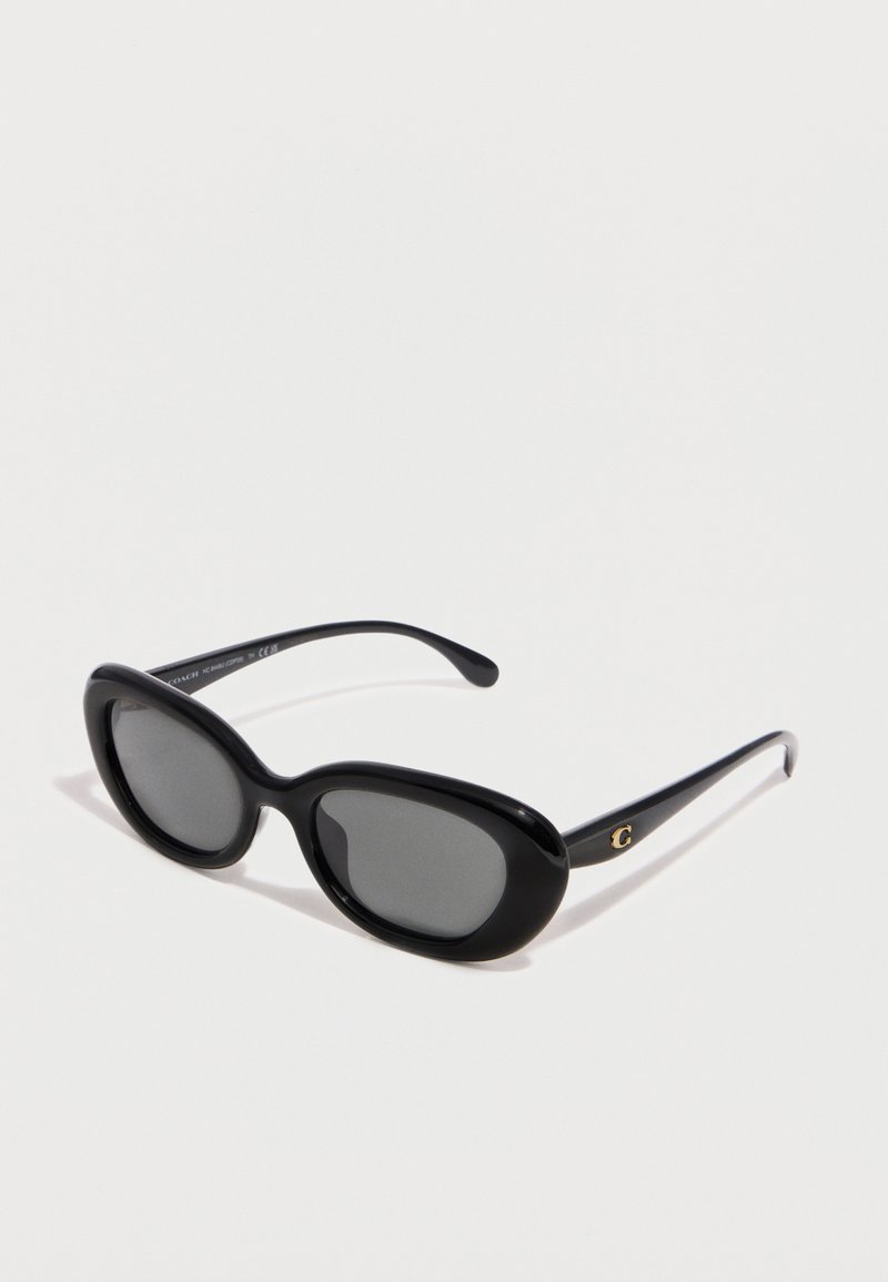 Μαύρα οβάλ γυαλιά ηλίου με σκουρόχρωμους φακούς και χρυσό λογότυπο "C" στο πλαϊνό μέρος, απεικονίζονται πάνω σε απλό λευκό φόντο.