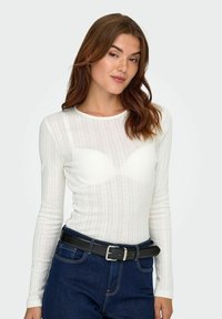 Vit långärmad topp med en ribbad textur och vertikala ränder samt en figursydd design. Matchad med mörkblå denim och ett svart bälte.