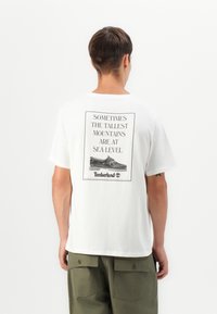 White cotton t-shirt with a rear graphic featuring black text and an image of shoes. The design includes a framed quote and brand logo.