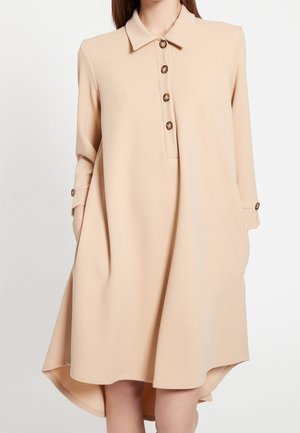 Kobieta w beżowej sukience z długim rękawem, kołnierzykiem, czterema guzikami z przodu i kieszeniami, stojąca na jednolitym tle.