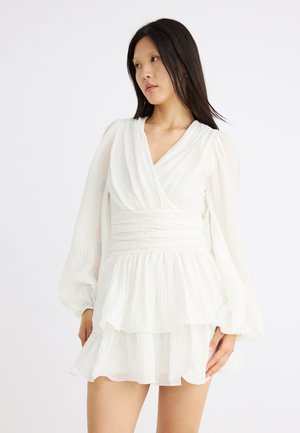 Moteris su ilgais tamsiais plaukais, dėvinti balta, ilgomis rankovėmis, sluoksniuota mini suknelę su raukšlėtu liemeniu, šiek tiek žiūrinti šonu prieš baltą foną.