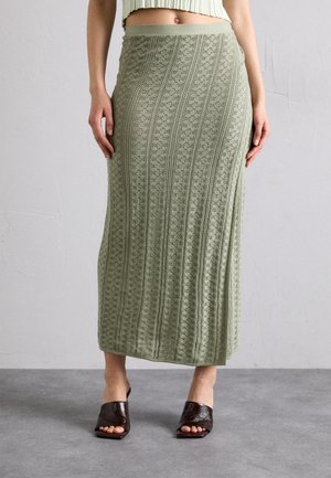 Mujer con falda midi de punto texturizada verde con una abertura, combinada con sandalias de tacón abiertas en marrón oscuro, de pie sobre un suelo gris.