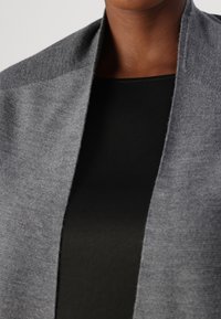 Cardigan gris de punto con un acabado texturizado y abertura frontal, superpuesto a una blusa negra, que muestra un diseño simple y contemporáneo.