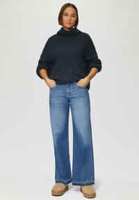 Navy gebreide trui met hoge kraag, gecombineerd met lichtblauwe wijde jeans en tan kleurige pantoffels. Eenvoudig ontwerp met geribbelde textuur.