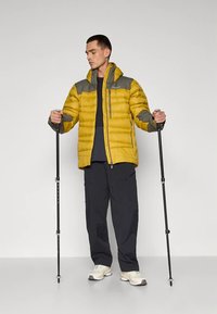 Geltona pūkinė striukė su pilkomis detalėmis, kišenėmis su užtrauktukais ir gobtuvu. Modelis laiko žygių lazdas, dėvi tamsias kelnes ir šviesios spalvos batus.