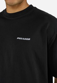 Muž v čiernom tričku s bielym logom "PEGADOR" umiestneným na strede hornej časti hrudníka, viditeľným od krku po stred trupu.