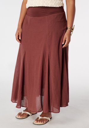Femme portant une longue jupe fluide marron, un haut crème texturé, un bracelet doré et des sandales blanches ornées de coquillages, debout sur un fond blanc.