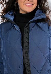 Leende person som bär hängande örhängen, svart polotröja och en blå vadderad jacka med uppvikt krage samt synlig dragkedja och knapp.